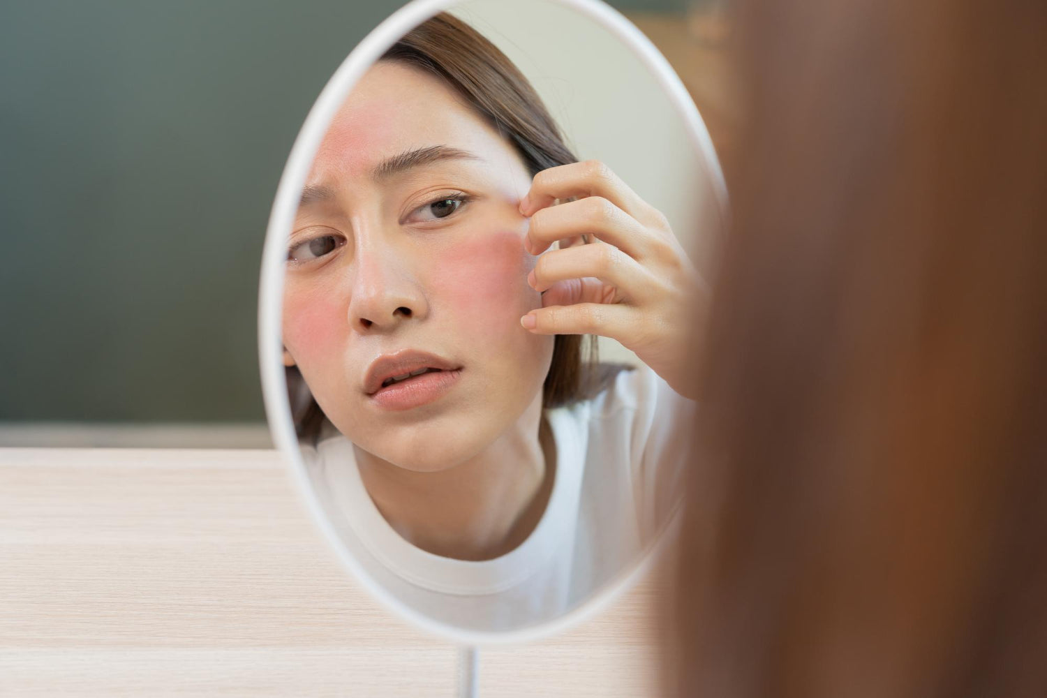 A woman looking in an oval mirror at severe facial redness, seeking the best cream for eczema itching relief.