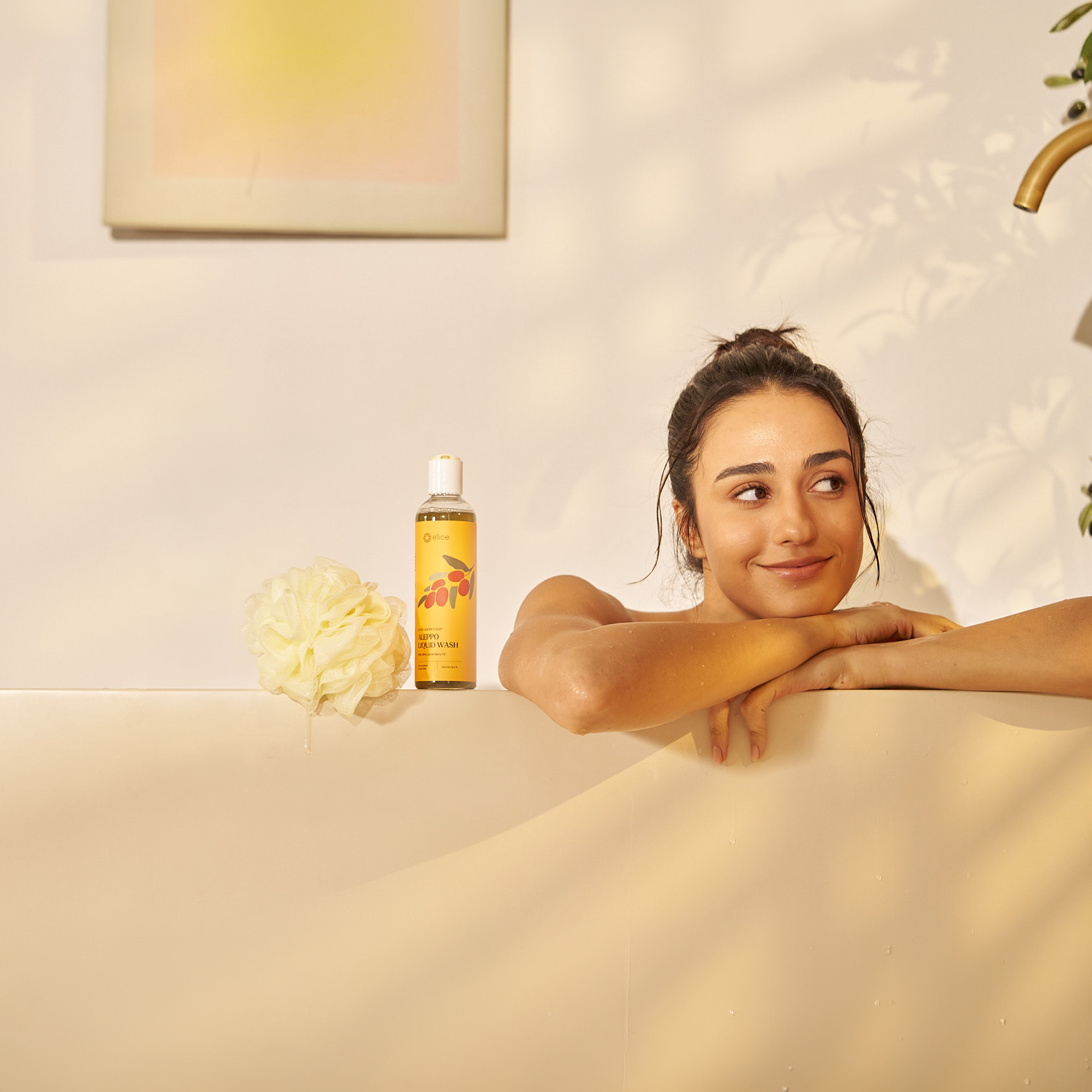 Woman relaxing in a bathtub with a bottle of e'lioe Aleppo liquid wash and foaming net