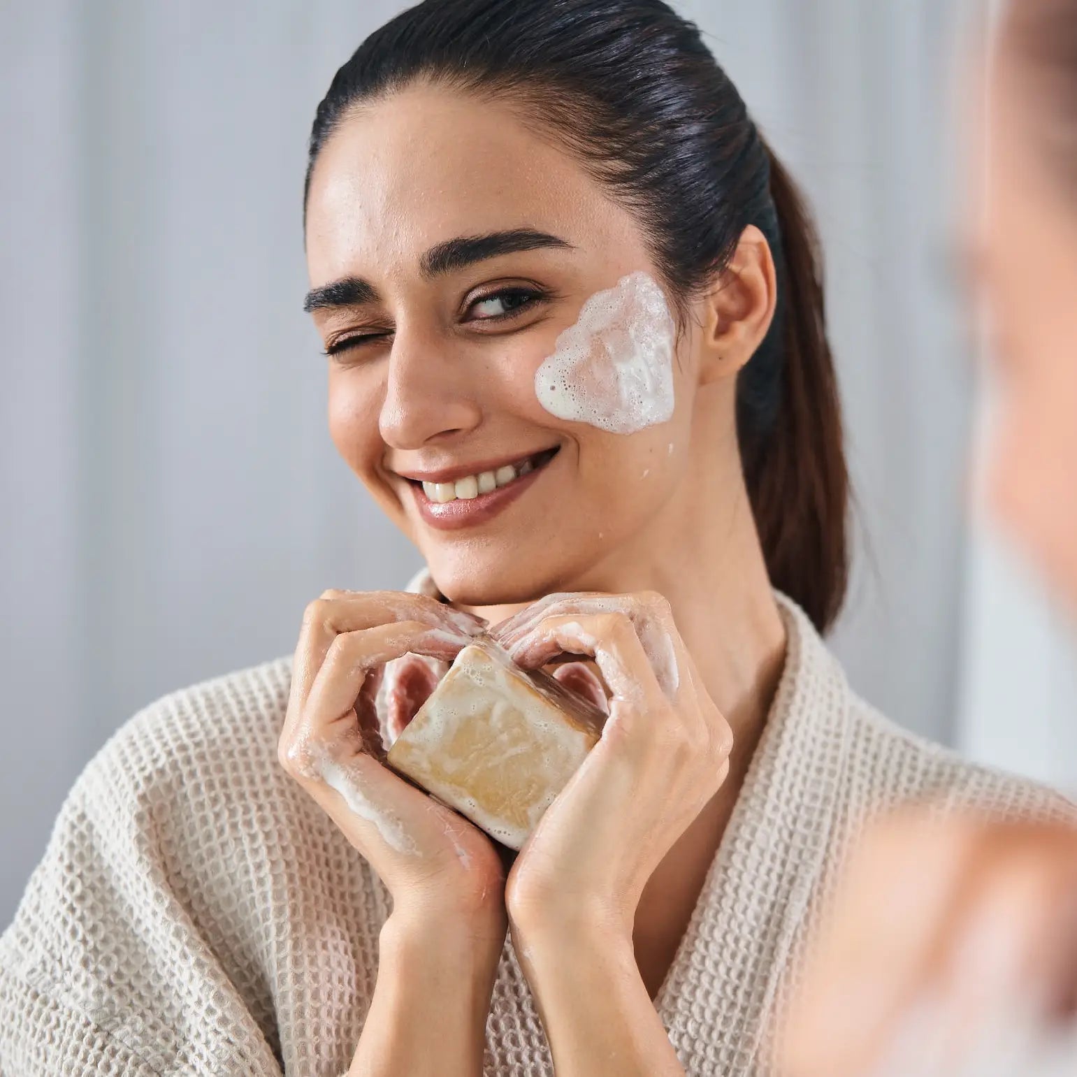 A smiling woman with lather on her cheek holding a gentle aleppo soap block, a natural cleanser for acne.